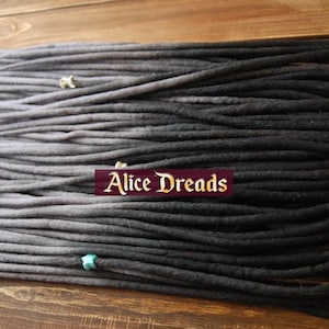 May include: A set of ombre grey and black synthetic dreadlocks. The dreadlocks are arranged in a neat pile on a wooden surface. The dreadlocks are approximately 50 cm long.