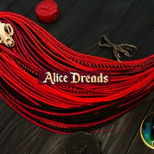May include: Red and black synthetic dreadlocks with a small white skull charm. The dreadlocks are arranged in a fan shape and are on a dark wooden surface. The text "Alice Dreads" is visible in the image.
