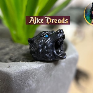 May include: A black bear head ring with blue eyes and an open mouth. The ring is set in a concrete planter with green foliage in the background. The text "Alice Dreads" is displayed on a maroon banner.
