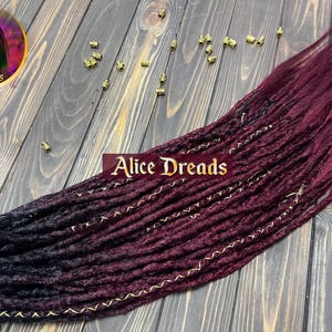 Synthetic dreads with black to burgundy ombre, available in 50-60 cm or 75-80 cm lengths. Features loose or dreaded ends with black and gold wrapped accents. Double Ended (DE) or Single Ended (SE) options.