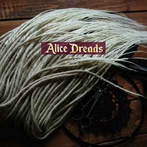Puede incluir: Un paquete de rastas sintéticas de color rubio claro, con algunas hebras blancas, dispuestas sobre una superficie de madera. Las rastas están atadas juntas con una cinta negra. El texto "Alice Dreads" es visible en la imagen.