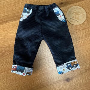 May include: Black corduroy trousers featuring an elasticated waistband and two front pockets lined with a white fabric printed with colourful construction vehicles. The trouser legs are cuffed at the bottom, revealing the same white construction vehicle print.