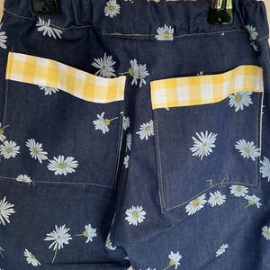 May include: Close-up of denim pants with a daisy floral pattern. The back pockets have yellow and white gingham trim. The pants are hanging from a metal hanger.