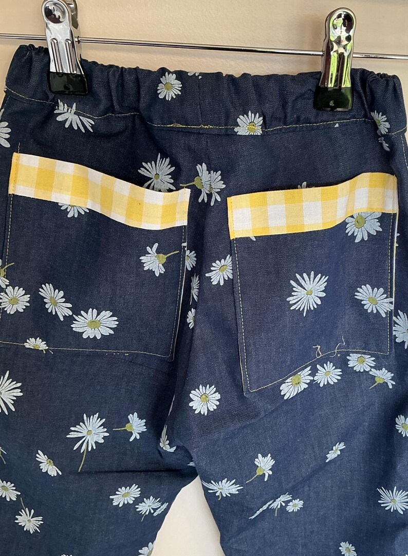 May include: Close-up of denim pants with a daisy floral pattern. The back pockets have yellow and white gingham trim. The pants are hanging from a metal hanger.