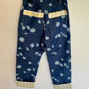 May include: Dark blue denim pants with a daisy floral pattern. The pants have yellow and white gingham accents on the pockets and cuffs. The pants are hanging on a metal hanger.
