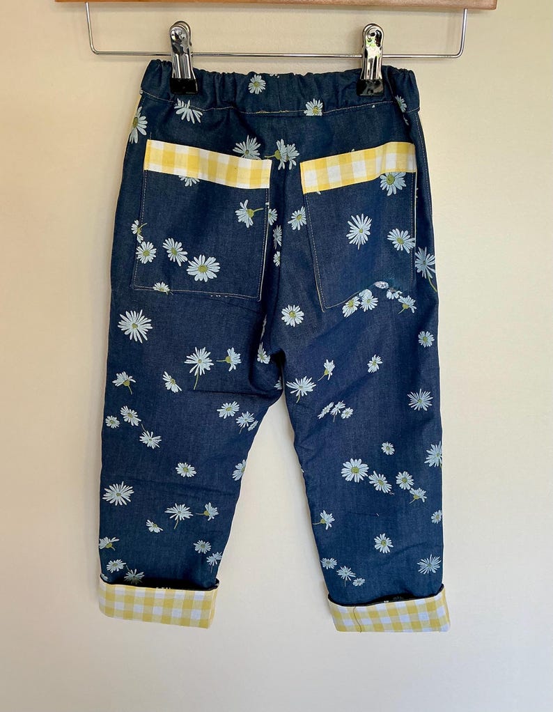 May include: Dark blue denim pants with a daisy floral pattern. The pants have yellow and white gingham accents on the pockets and cuffs. The pants are hanging on a metal hanger.