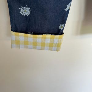 May include: Dark blue fabric with a white daisy pattern, cuffed to reveal a yellow and white gingham lining. The cuff is folded up, showcasing the contrasting pattern. The fabric is likely denim or a similar material.