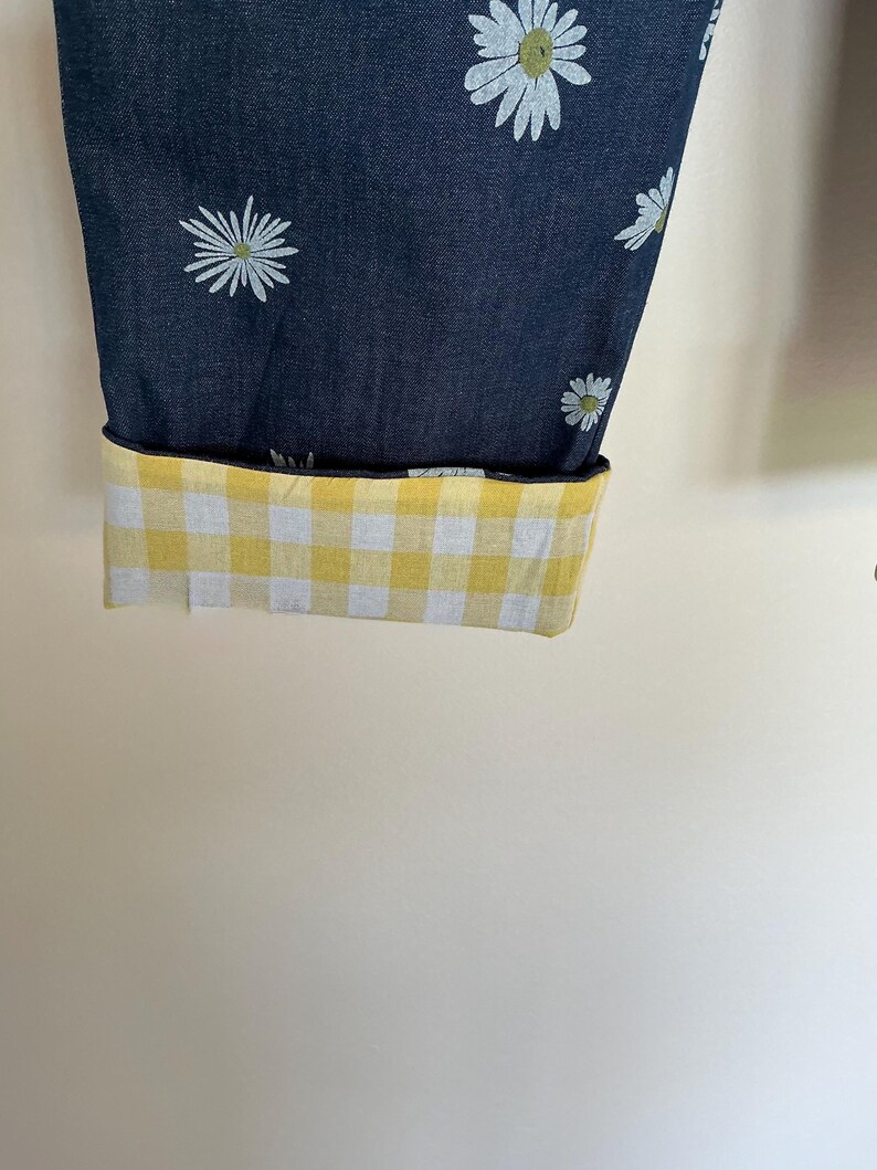 May include: Dark blue fabric with a white daisy pattern, cuffed to reveal a yellow and white gingham lining. The cuff is folded up, showcasing the contrasting pattern. The fabric is likely denim or a similar material.
