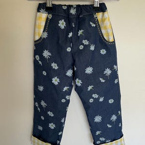 May include: Blue denim pants with a white daisy print. The pockets and cuffs are lined with yellow and white gingham fabric. The pants are hanging on a wooden hanger.