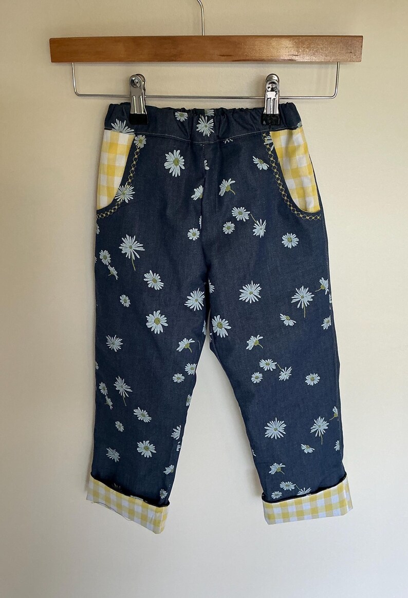 May include: Blue denim pants with a white daisy print. The pockets and cuffs are lined with yellow and white gingham fabric. The pants are hanging on a wooden hanger.