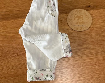 Può includere: Pantaloni bianchi a coste con stampa floreale e di conigli sulla fodera delle tasche e sui risvolti. Un'etichetta rotonda in legno con l'illustrazione di una volpe e la scritta Stitched by Chris è posizionata accanto ai pantaloni.