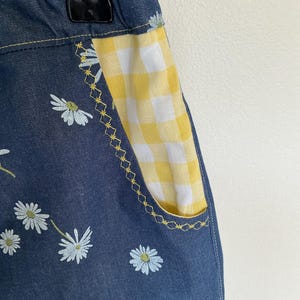 May include: Close-up of denim shorts with a daisy floral pattern. The pocket is yellow and white gingham with yellow stitching. The shorts are hanging on a metal hanger.