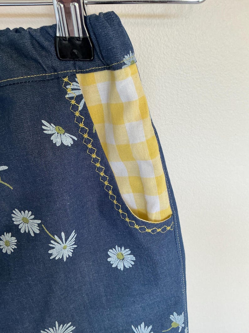 May include: Close-up of denim shorts with a daisy floral pattern. The pocket is yellow and white gingham with yellow stitching. The shorts are hanging on a metal hanger.