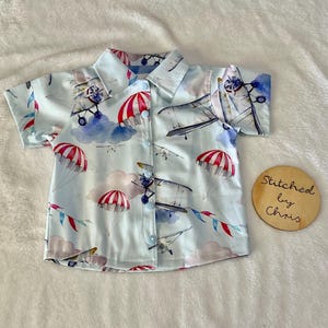 May include: Pale blue short-sleeved shirt featuring a pattern of aeroplanes, parachutes, and clouds. The shirt has a button-down front and a collar. The design includes red, white, and blue accents. The shirt is made of a soft, lightweight fabric. The text "Stitched by Chris" is on a wooden circle.