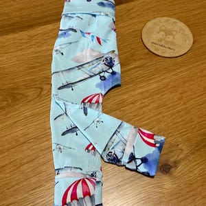 May include: Light blue trousers featuring a repeating pattern of vintage aeroplanes, white clouds, and red and white striped parachutes. The trousers have a folded waistband and cuffs at the bottom of the legs, with a smooth fabric finish throughout.