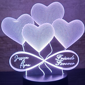 Puede incluir: Un símbolo de infinito blanco impreso en 3D con cuatro globos en forma de corazón unidos. El símbolo de infinito tiene el texto "Jessica & Ryan" en un lado y "Friends Forever" en el otro. La lámpara tiene un mando a distancia con múltiples luces de colores.