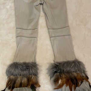 Puede incluir: Pantalones vaqueros de color gris claro con canesú trasero, dos bolsillos traseros con remaches metálicos y costuras horizontales sobre las rodillas. Las perneras terminan en puños superpuestos de piel sintética gris y plumas marrones.
