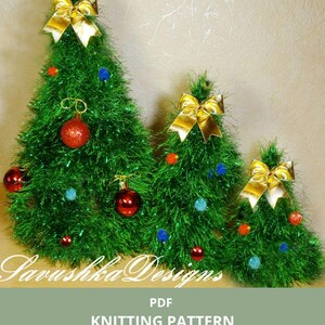 Könnte beinhalten: Drei grüne Weihnachtsbaumdekorationen in verschiedenen Größen, verziert mit goldenen Schleifen, roten Kugeln und bunten Pompons. Der Text weist auf ein Strickmuster für die Bäume hin.