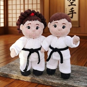 May include: Two knitted dolls dressed in white martial arts uniforms with black belts. The doll on the left has brown curly hair with a red accent, while the doll on the right has short brown hair. Both dolls feature embroidered facial details.