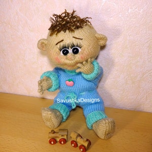 A knitted baby doll wearing a blue jumpsuit and a hat, sitting in a woven basket with a pillow. The doll is handmade, with a cute face, black eyes, and an adorable expression. The PDF pattern is available for download and includes detailed instructions.