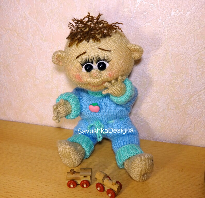 A knitted baby doll wearing a blue jumpsuit and a hat, sitting in a woven basket with a pillow. The doll is handmade, with a cute face, black eyes, and an adorable expression. The PDF pattern is available for download and includes detailed instructions.
