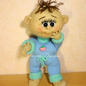 A knitted baby doll wearing a blue jumpsuit and a hat, sitting in a woven basket with a pillow. The doll is handmade, with a cute face, black eyes, and an adorable expression. The PDF pattern is available for download and includes detailed instructions.