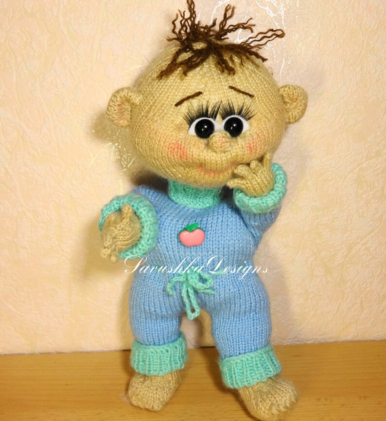 A knitted baby doll wearing a blue jumpsuit and a hat, sitting in a woven basket with a pillow. The doll is handmade, with a cute face, black eyes, and an adorable expression. The PDF pattern is available for download and includes detailed instructions.