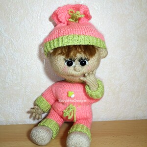 A knitted baby doll wearing a blue jumpsuit and a hat, sitting in a woven basket with a pillow. The doll is handmade, with a cute face, black eyes, and an adorable expression. The PDF pattern is available for download and includes detailed instructions.