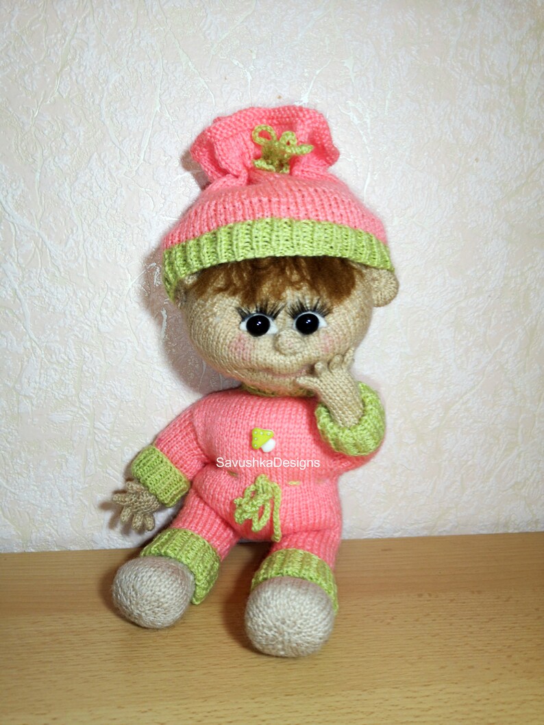 A knitted baby doll wearing a blue jumpsuit and a hat, sitting in a woven basket with a pillow. The doll is handmade, with a cute face, black eyes, and an adorable expression. The PDF pattern is available for download and includes detailed instructions.