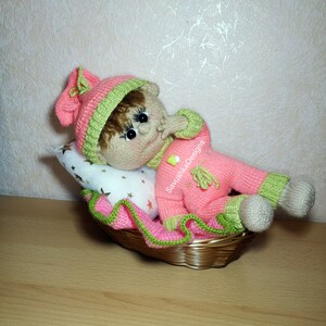 A knitted baby doll wearing a blue jumpsuit and a hat, sitting in a woven basket with a pillow. The doll is handmade, with a cute face, black eyes, and an adorable expression. The PDF pattern is available for download and includes detailed instructions.