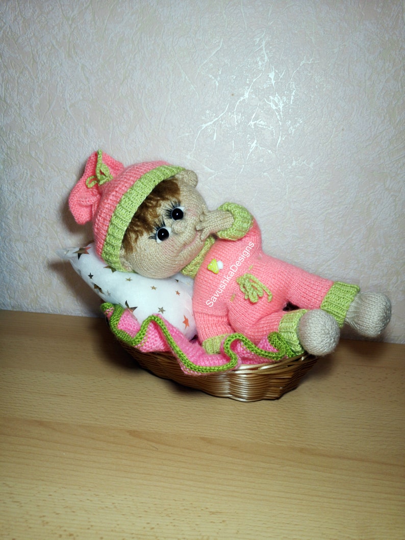 A knitted baby doll wearing a blue jumpsuit and a hat, sitting in a woven basket with a pillow. The doll is handmade, with a cute face, black eyes, and an adorable expression. The PDF pattern is available for download and includes detailed instructions.
