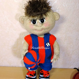 Könnte beinhalten: Eine gestrickte Puppe, die ein rot-blau gestreiftes Fußballtrikot und Shorts trägt und einen Fußball hält. Die Puppe hat braunes Haar und große Augen. Die Puppe steht auf einer Holzoberfläche. "Savushka Designs" steht auf dem Trikot.
