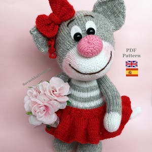 Puede incluir: Una muñeca de gato tejida a mano, de color gris con hocico blanco, nariz rosa y un lazo rojo en la cabeza. El gato lleva una falda roja y sostiene un ramo de rosas de color rosa claro. La imagen incluye el texto PDF Pattern y banderas de RU y España.
