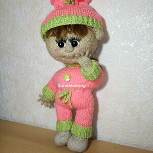 A knitted baby doll wearing a blue jumpsuit and a hat, sitting in a woven basket with a pillow. The doll is handmade, with a cute face, black eyes, and an adorable expression. The PDF pattern is available for download and includes detailed instructions.