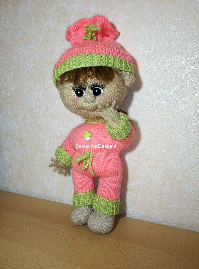 A knitted baby doll wearing a blue jumpsuit and a hat, sitting in a woven basket with a pillow. The doll is handmade, with a cute face, black eyes, and an adorable expression. The PDF pattern is available for download and includes detailed instructions.