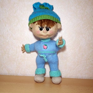 A knitted baby doll wearing a blue jumpsuit and a hat, sitting in a woven basket with a pillow. The doll is handmade, with a cute face, black eyes, and an adorable expression. The PDF pattern is available for download and includes detailed instructions.