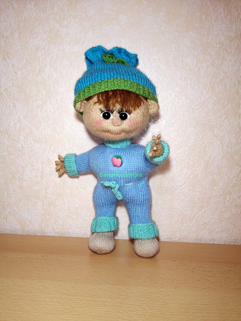 A knitted baby doll wearing a blue jumpsuit and a hat, sitting in a woven basket with a pillow. The doll is handmade, with a cute face, black eyes, and an adorable expression. The PDF pattern is available for download and includes detailed instructions.