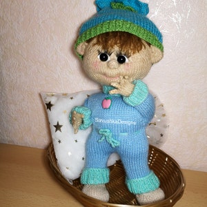 A knitted baby doll wearing a blue jumpsuit and a hat, sitting in a woven basket with a pillow. The doll is handmade, with a cute face, black eyes, and an adorable expression. The PDF pattern is available for download and includes detailed instructions.