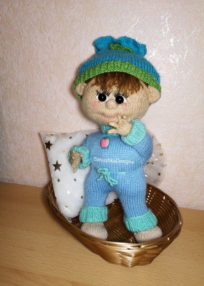 A knitted baby doll wearing a blue jumpsuit and a hat, sitting in a woven basket with a pillow. The doll is handmade, with a cute face, black eyes, and an adorable expression. The PDF pattern is available for download and includes detailed instructions.