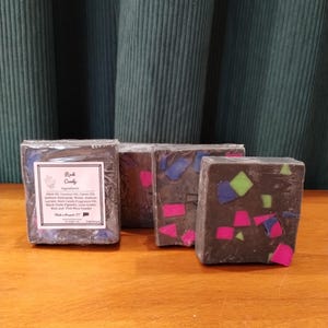 May include: Three rectangular bars of dark gray soap wrapped in clear plastic. The soap features embedded geometric shards in bright neon pink, lime green, and blue. One bar has a white label with the text Rock Candy and a list of ingredients.