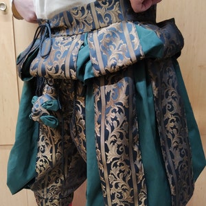 May include: Green and black Renaissance-style breeches with gold floral brocade accents. The breeches have a high waist and are gathered at the knee. A small pouch hangs from the waist. The person is wearing red socks.
