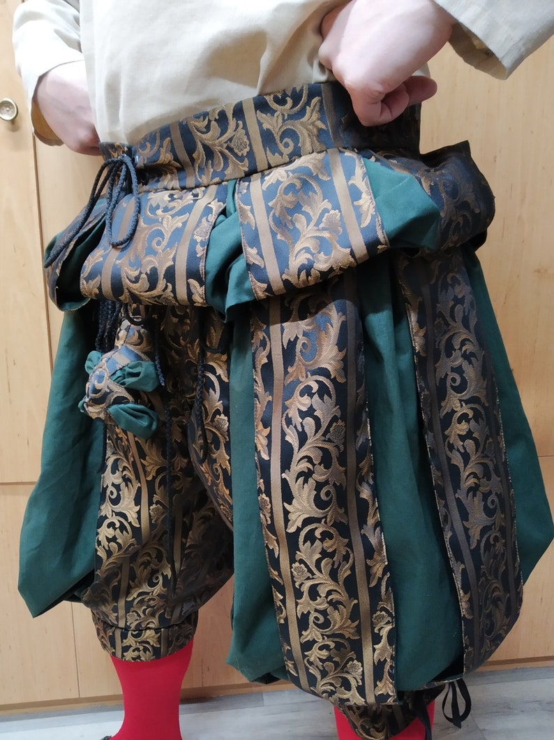 May include: Green and black Renaissance-style breeches with gold floral brocade accents. The breeches have a high waist and are gathered at the knee. A small pouch hangs from the waist. The person is wearing red socks.