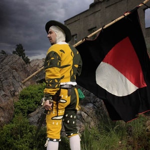 Könnte beinhalten: Ein Mann in einem gelben und grünen gemusterten Kostüm hält eine schwarze Flagge mit einem roten und weißen Kreis in der Mitte. Er steht vor einem steinernen Gebäude mit einem bewölkten Himmel im Hintergrund.