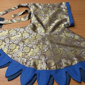 May include: A gold and brown patterned fabric garment with a blue scalloped edge. The fabric features a repeating design of animals and floral elements. The garment has a matching tie and a blue trim.