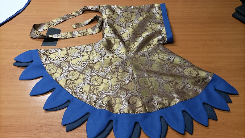 May include: A gold and brown patterned fabric garment with a blue scalloped edge. The fabric features a repeating design of animals and floral elements. The garment has a matching tie and a blue trim.