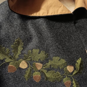 Op de afbeelding: Een donkergrijze capuchonmantel met een bruine voering en een geborduurd ontwerp van groene eikenbladeren en bruine eikels.