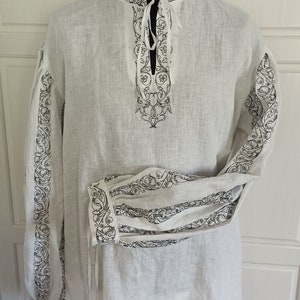 May include: A white linen shirt with black embroidered floral designs on the collar, cuffs, and front. The shirt has a tie at the neck.