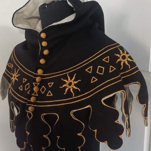 May include: Black hooded cloak with gold embroidered sun and geometric designs. The cloak has a scalloped edge and is fastened with six gold buttons. The hood lining is a light beige color.
