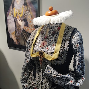 May include: A black and white lace dress with yellow ribbon and red and blue embellishments. The dress is on a mannequin with a white lace collar and a wooden stand.
