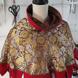 May include: A decorative cape with a brown and gold patterned fabric featuring animal and plant motifs. The cape has a red hood and a scalloped red trim along the bottom edge. The cape is displayed on a mannequin.
